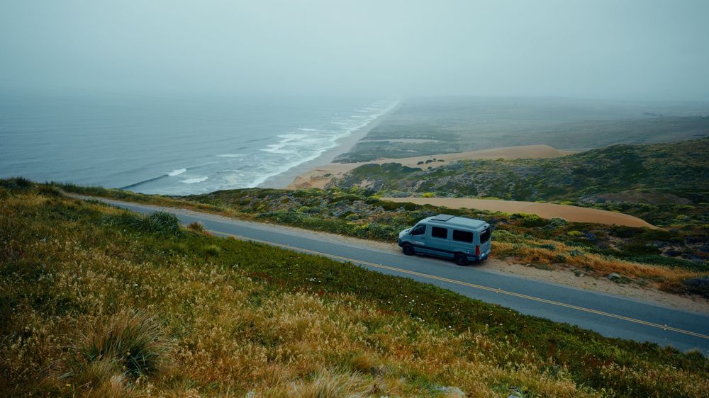 Camper van driving along misty coastal road with ocean views.
