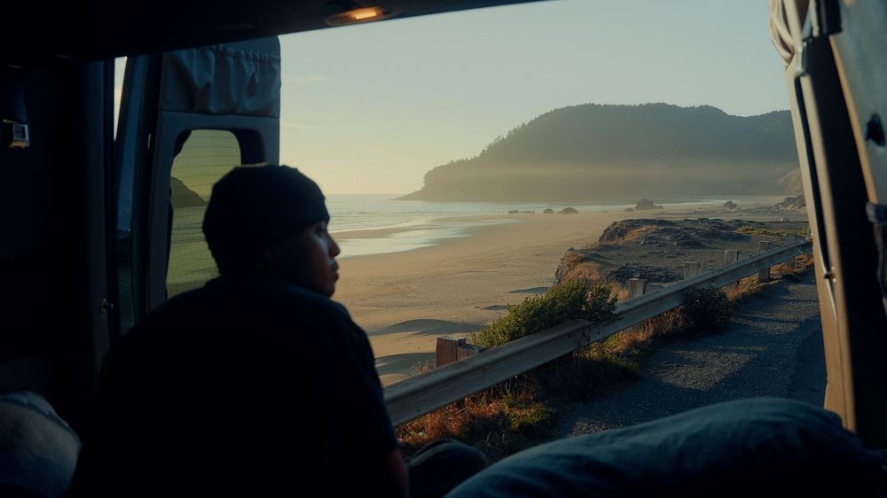 Coastal view through van window showing beach and mountains at sunset.