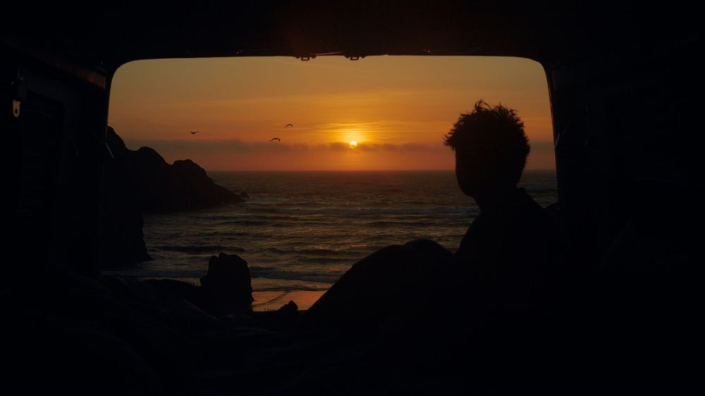 Silhouette view of sunset over ocean through camper van window during golden hour.