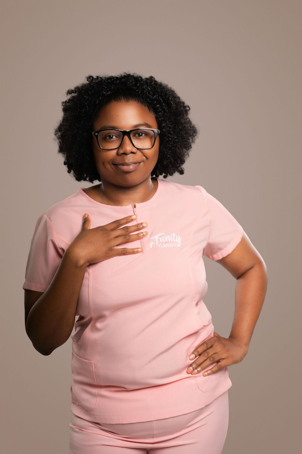 Medical professional in pink uniform and glasses poses with hand on heart.