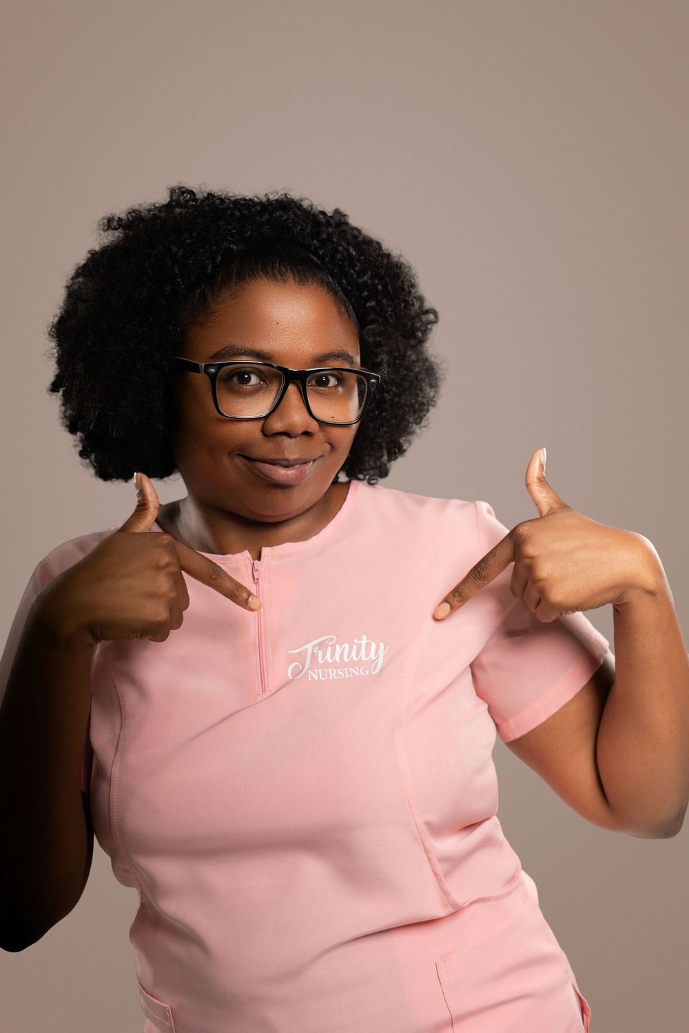 Healthcare worker in pink scrubs and glasses points to logo on uniform with enthusiasm.