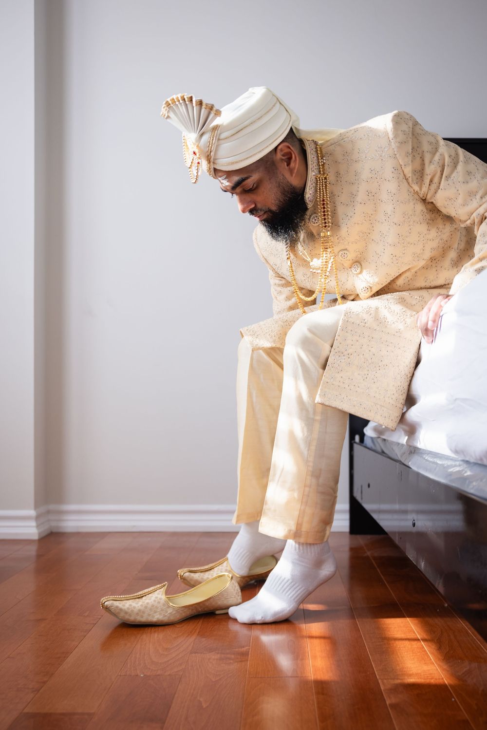 A groom in traditional cream-colored wedding attire sits on a bed while putting on gold mojari shoes.