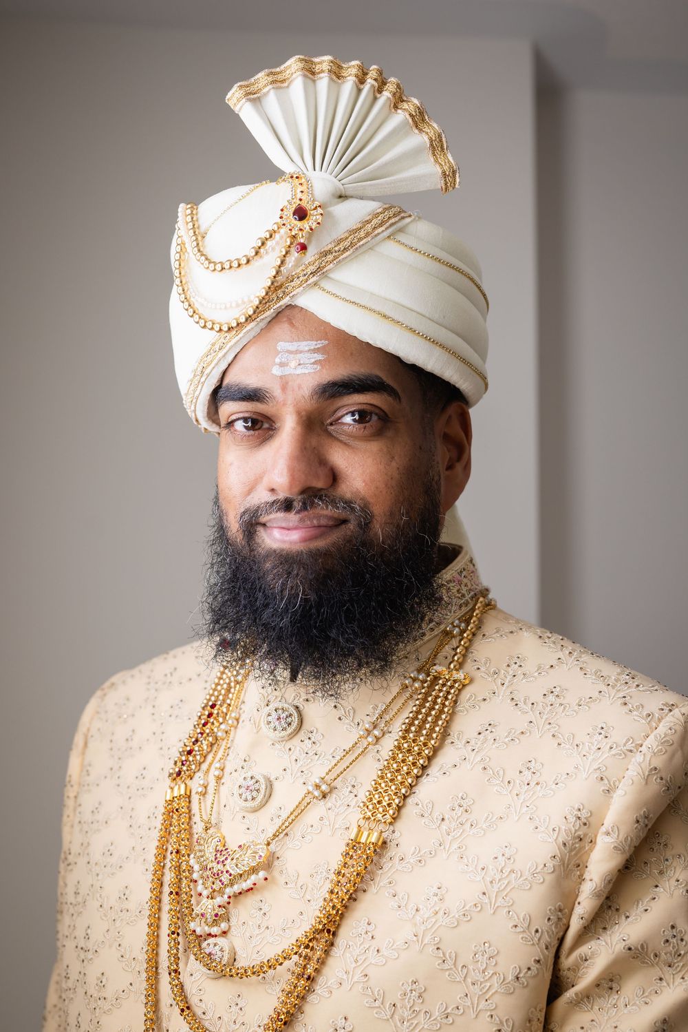 A groom in traditional Indian wedding attire wearing a cream sherwani, gold jewelry and white turban with gold accents.