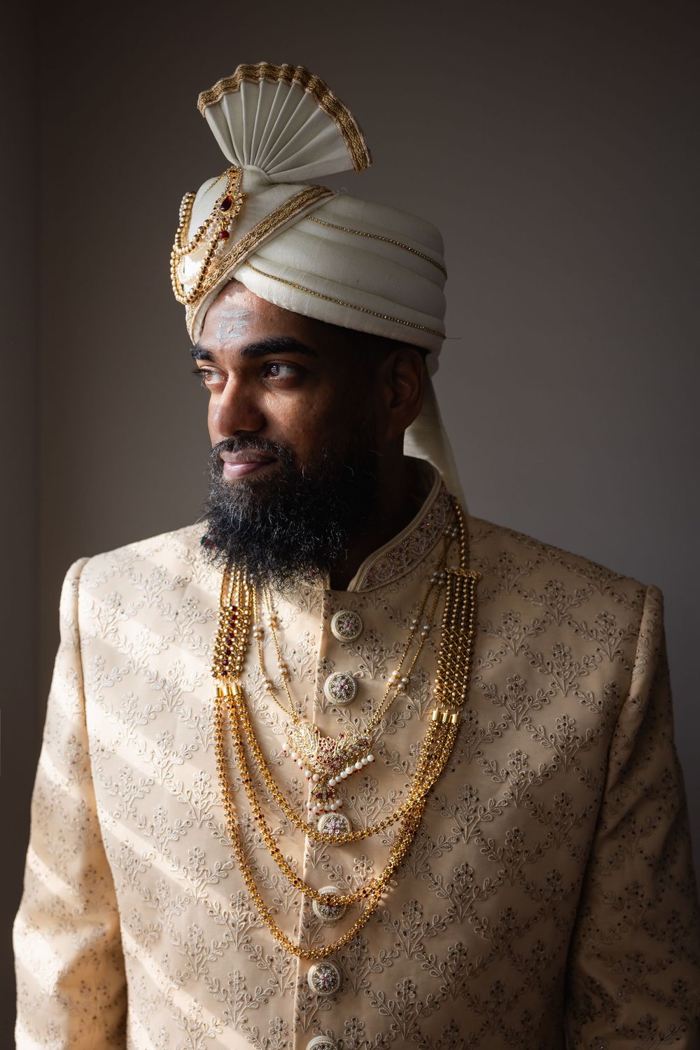 Traditional Indian groom wearing an elegant cream sherwani with gold necklaces and a white turban in a studio portrait series.