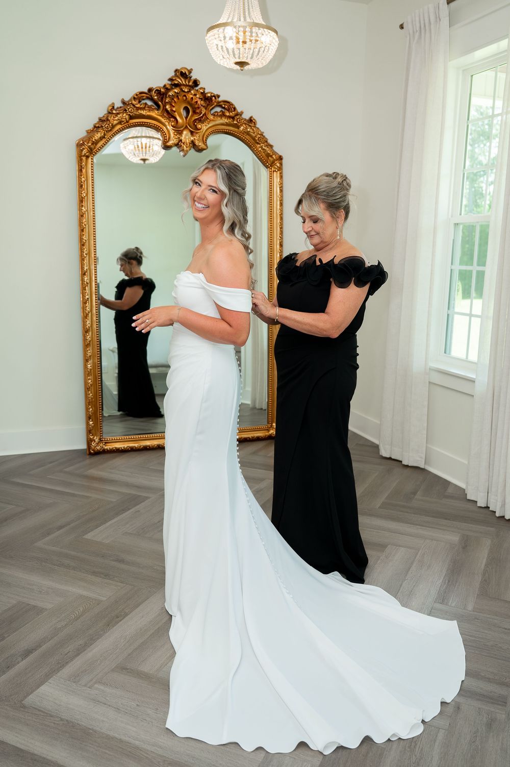 Elegant wedding dress fitting scene with ornate gold mirror and crystal chandelier.