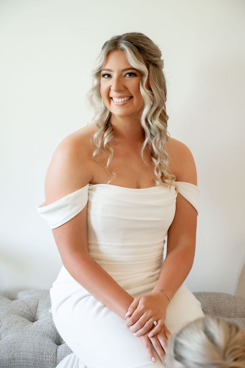 A bride in a white off-shoulder dress poses against a light background during her wedding preparation.