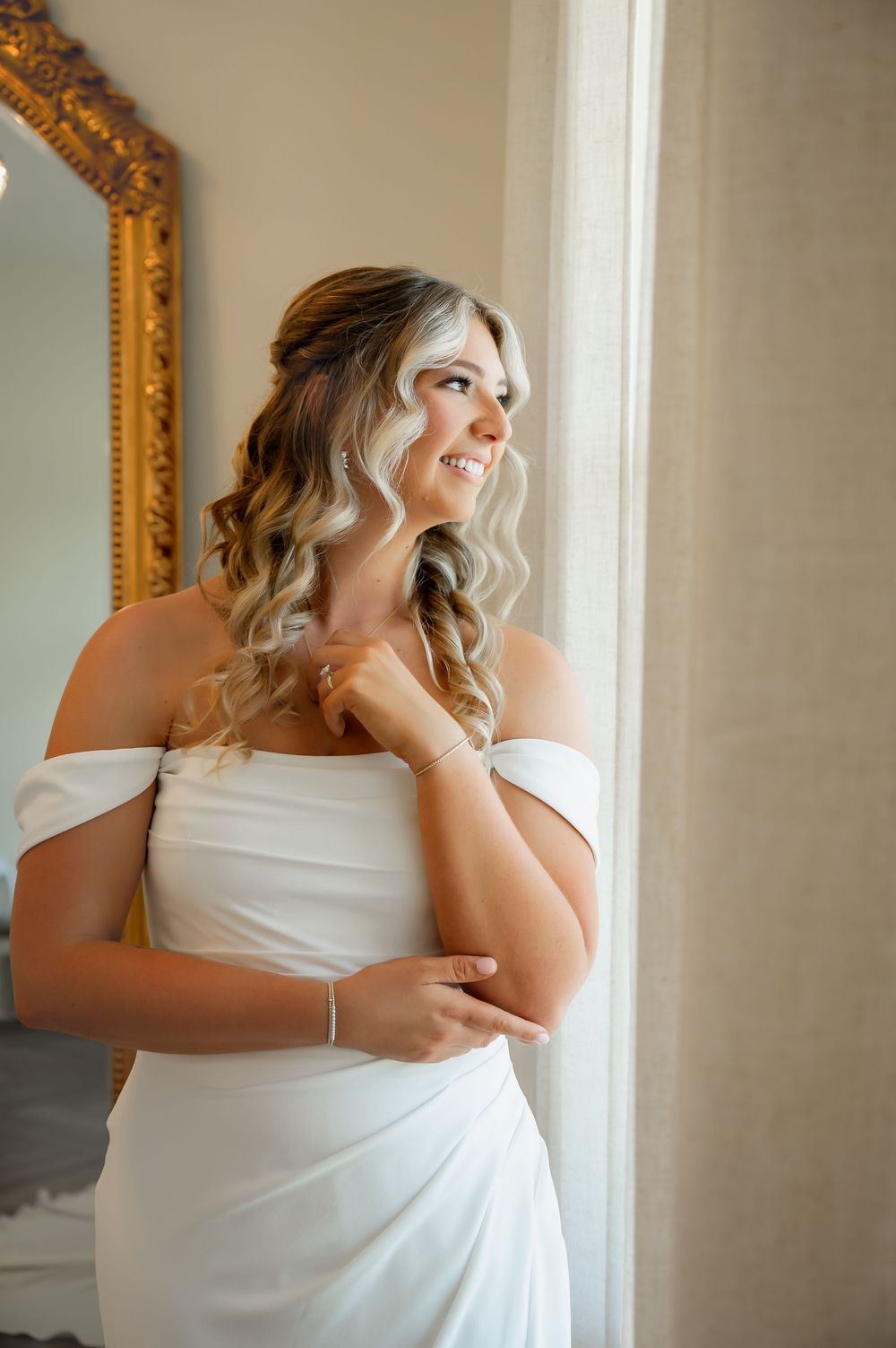 A bride in a white gown poses elegantly near an ornate gold-framed mirror.