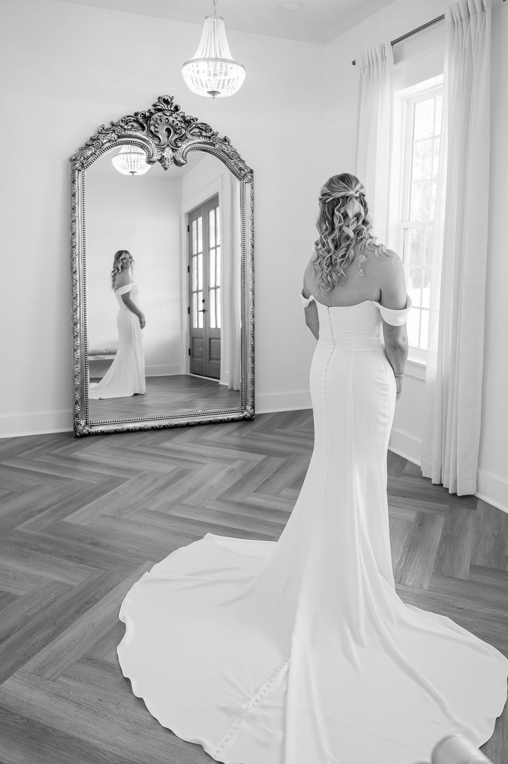 A flowing white wedding dress is showcased in a bright room with herringbone floors and ornate mirrors.