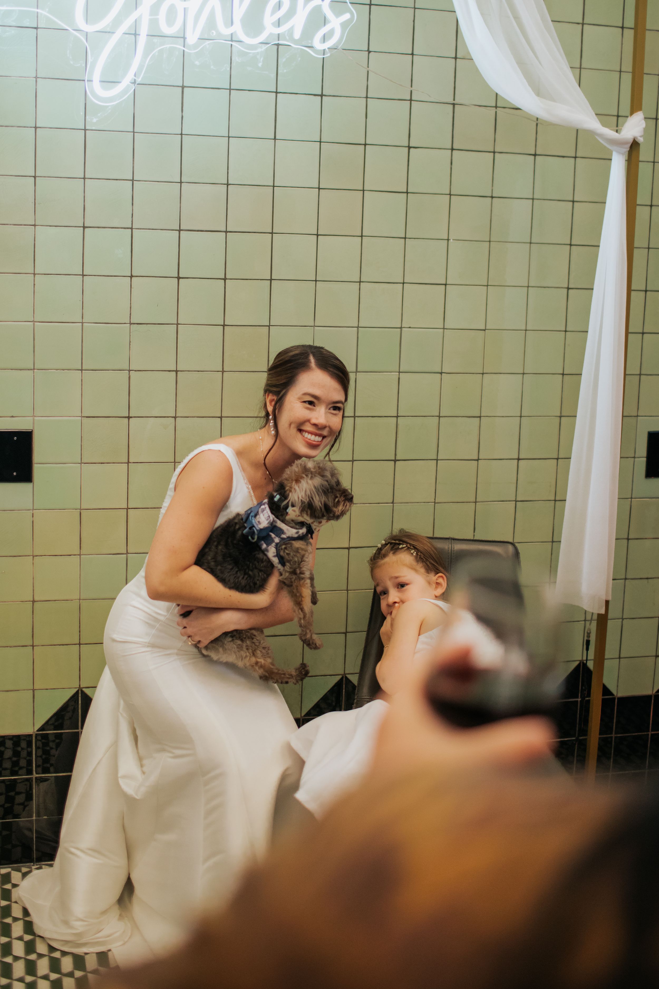 A person in white attire sits on a tiled bathroom floor with a small dog during a wedding event.