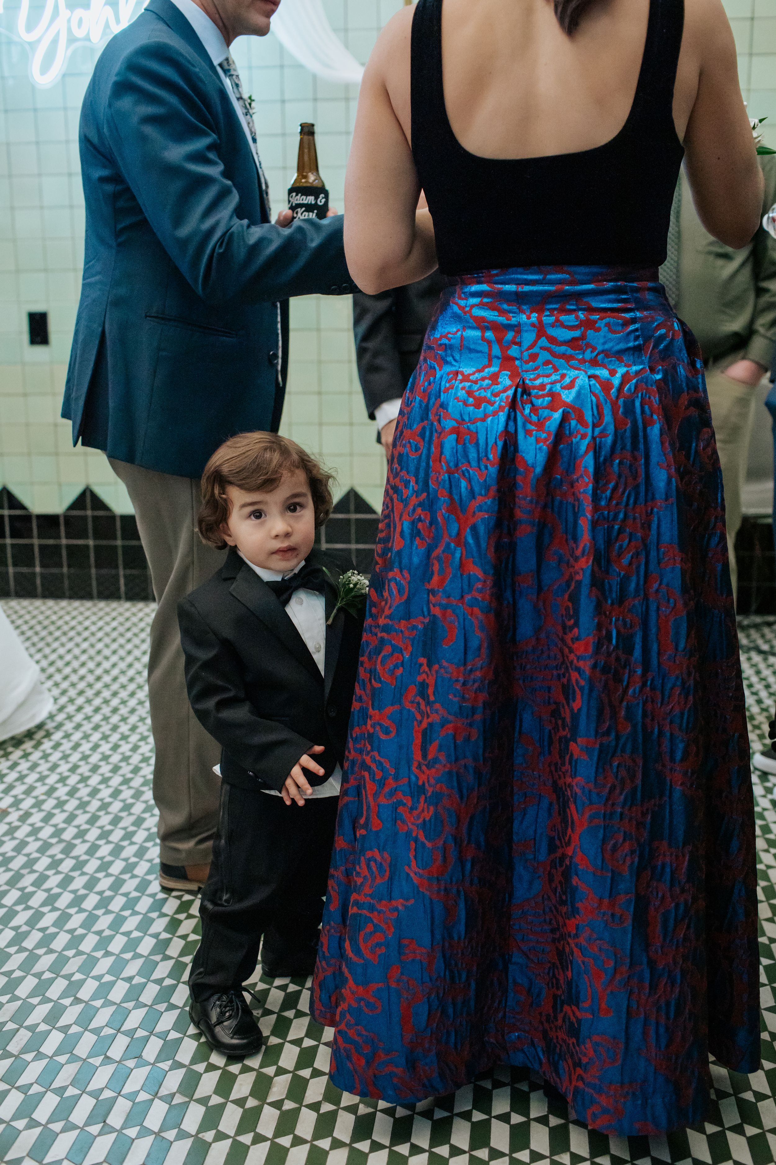 A young child in formal wear stands next to someone wearing a blue and red patterned skirt at an event.