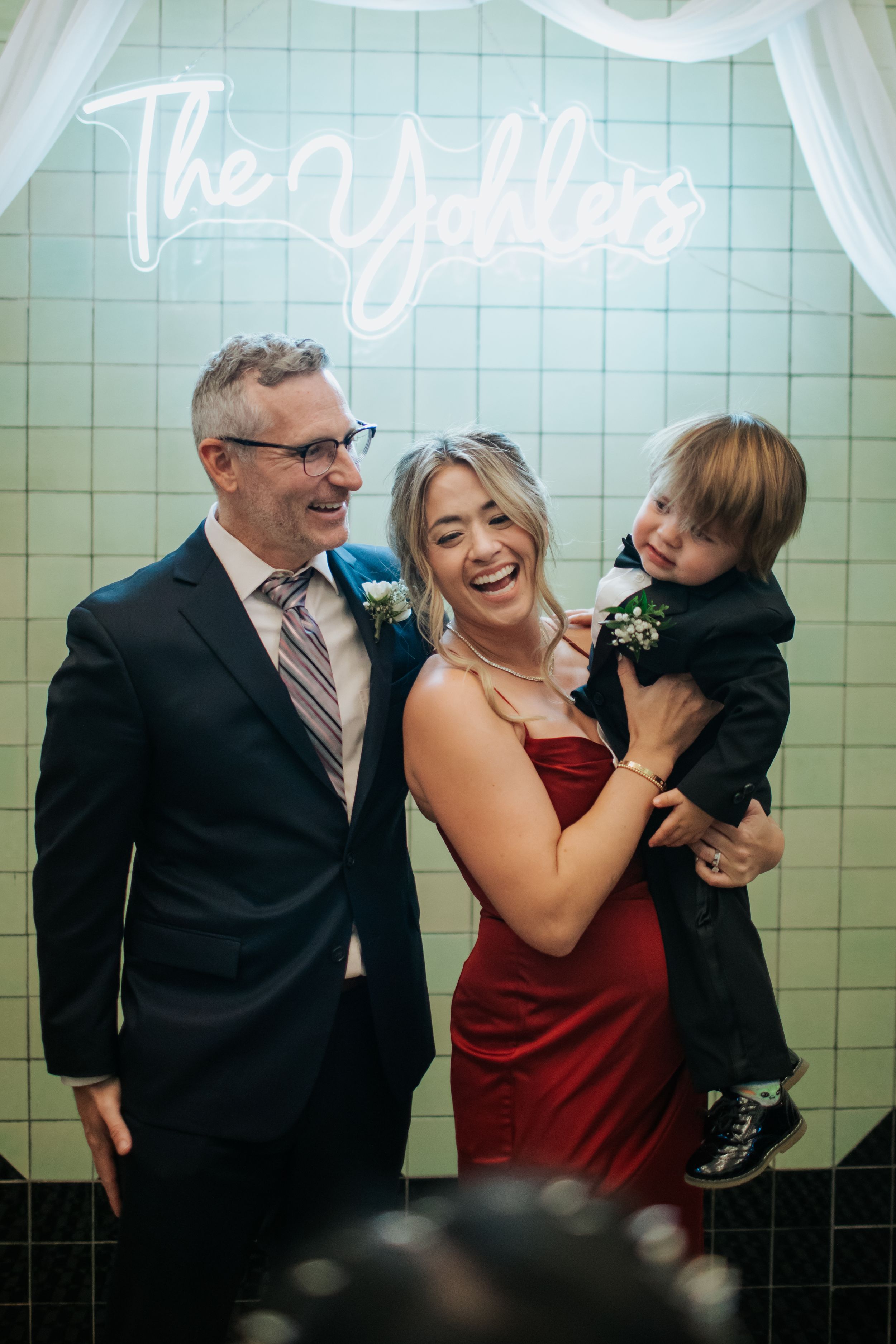 Family poses beneath neon wedding sign against mint green tile wall.