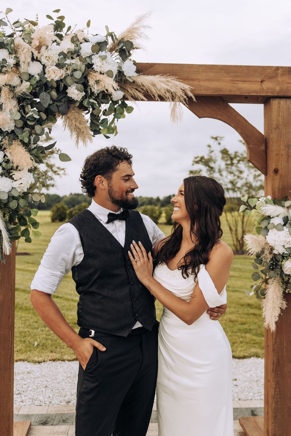 Smiling bride and groom pose under a wooden arch adorned with white roses and pampas grass outdoors.