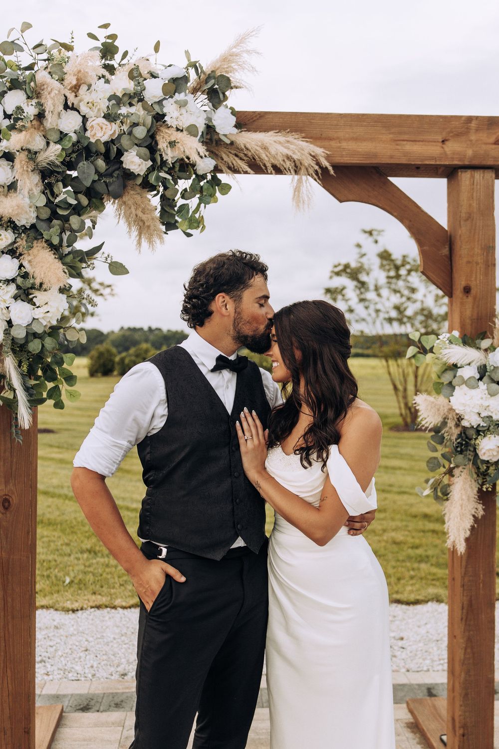Groom leans in to kiss bride under a rustic wooden arch decorated with white florals and pampas grass.