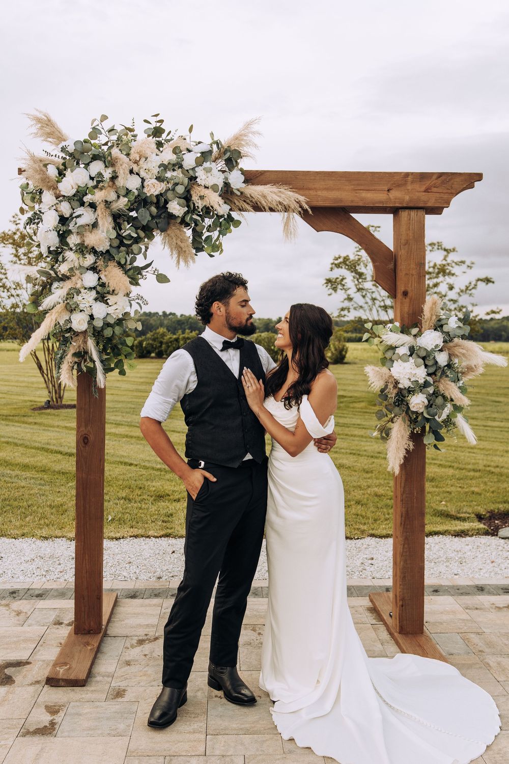Bride and groom kiss beneath a wooden wedding arch with white roses and pampas grass on a green lawn.