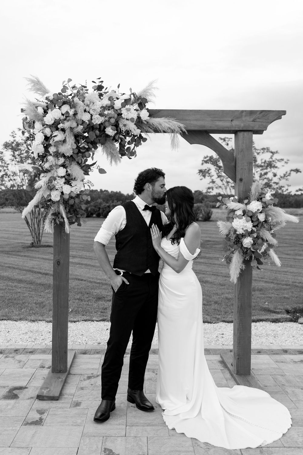 Black and white photo of bride and groom kissing under a floral wooden arch with pampas grass accents.