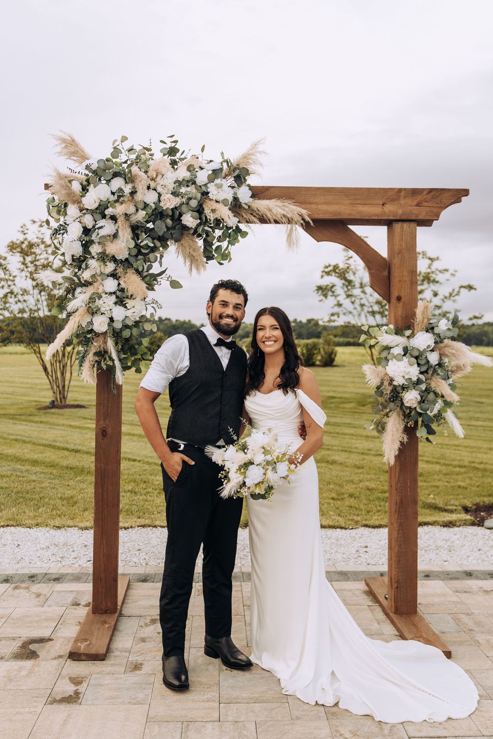 Bride and groom smile together under a rustic wooden arch with white roses and pampas grass outdoors.