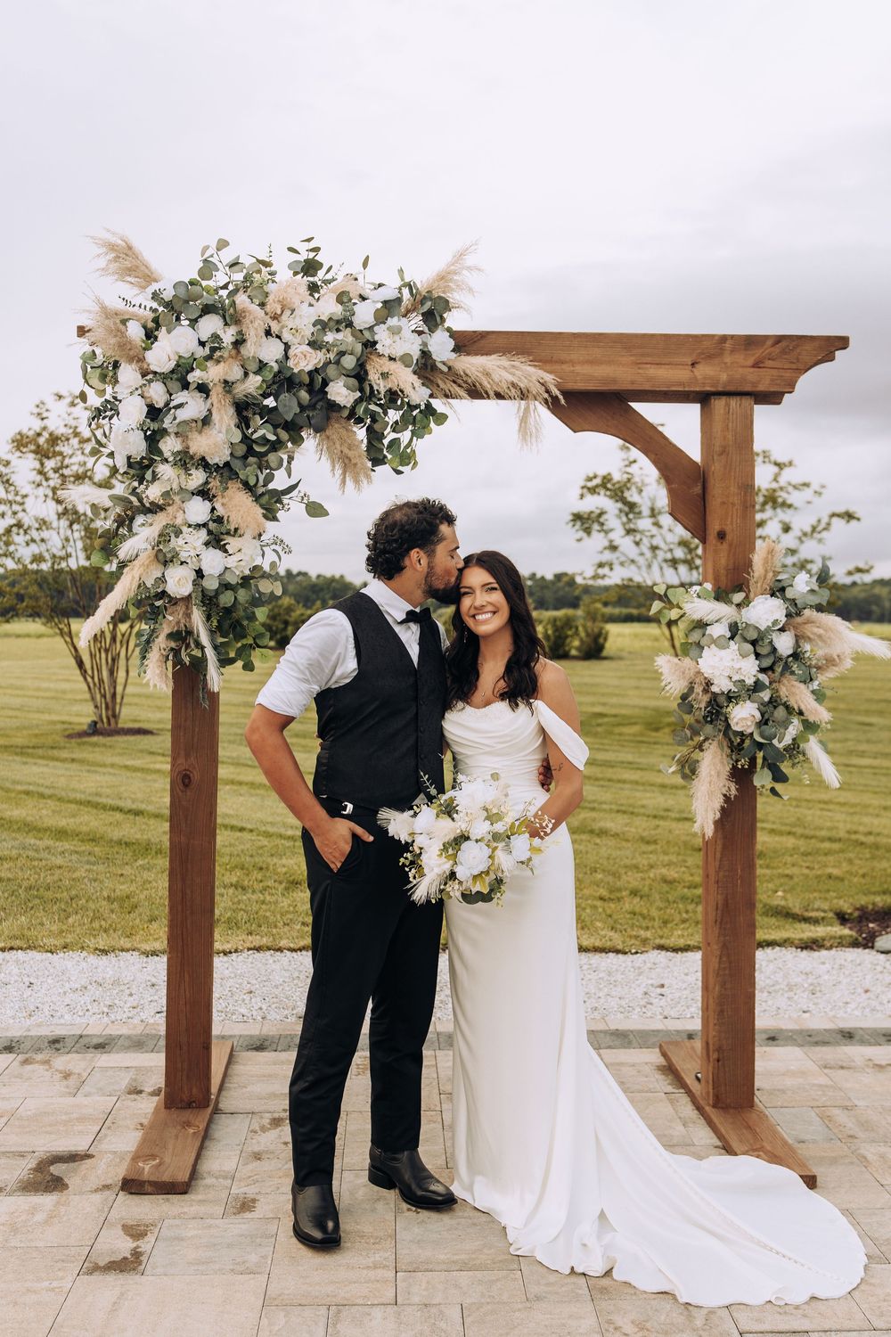 Groom kisses bride on the cheek as she smiles holding a white bouquet under a wooden floral arch.