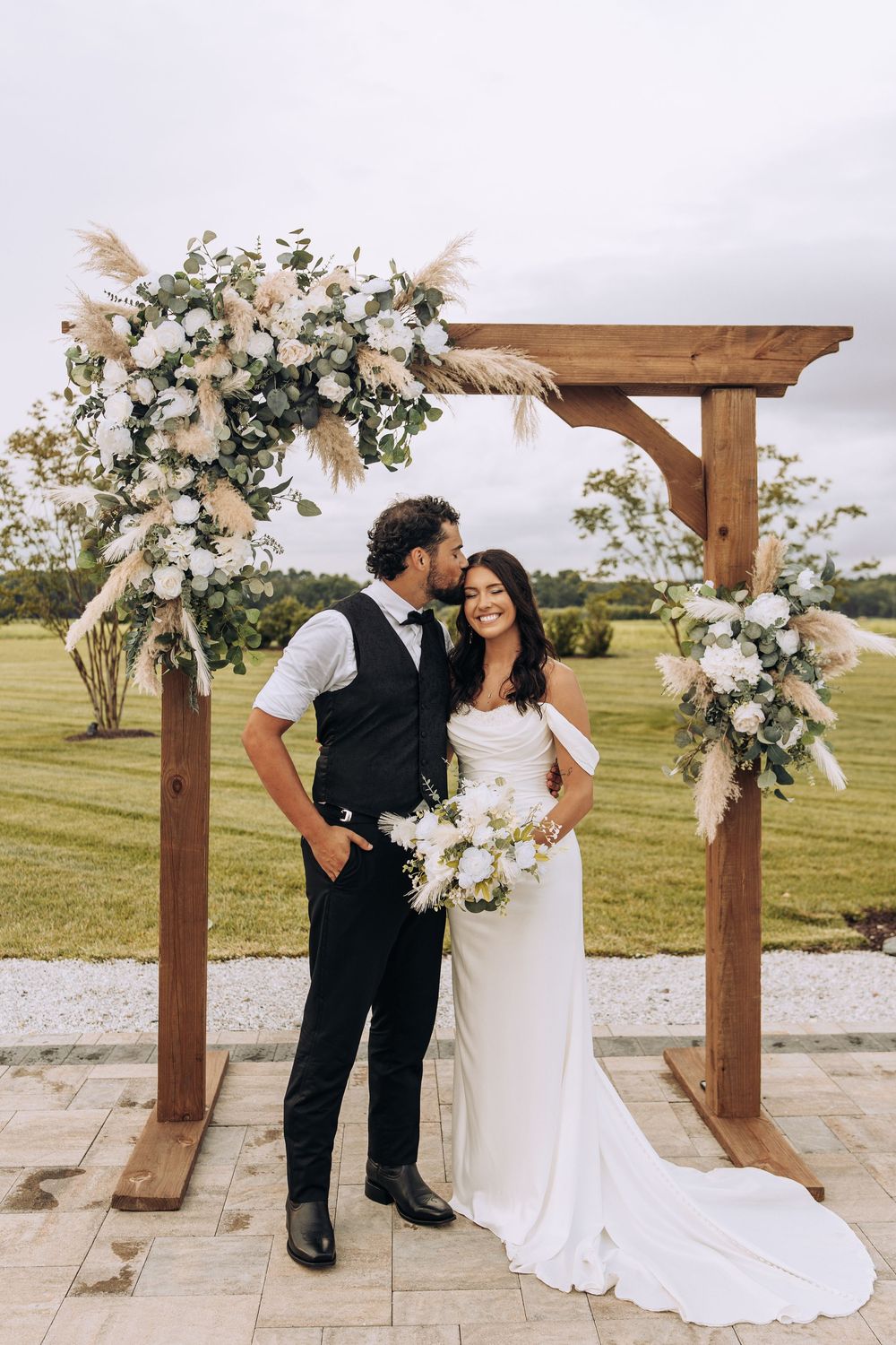 Groom kisses smiling bride on the cheek under a wooden arch with white roses and pampas grass decor.