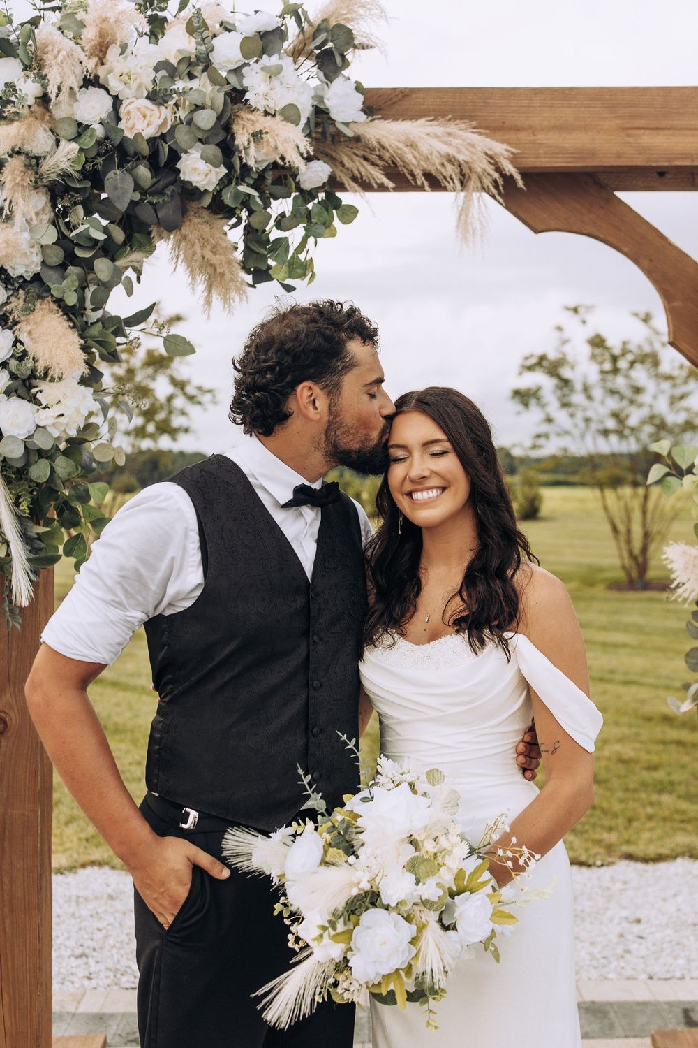 Groom kisses bride on the cheek as she smiles holding a white bouquet beneath a rustic floral arch.