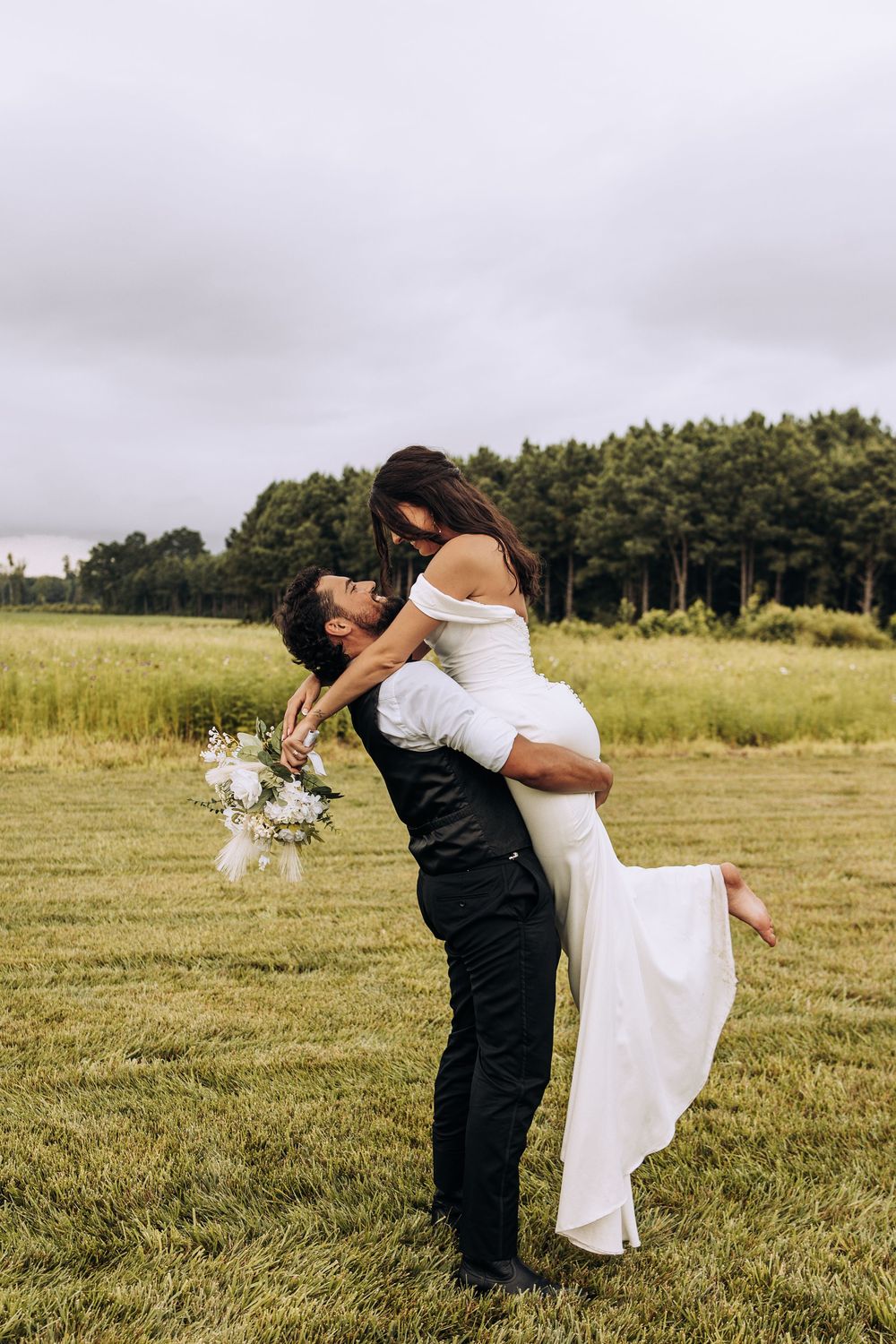 Groom lifts bride in white off-shoulder gown while holding bouquet in green field.