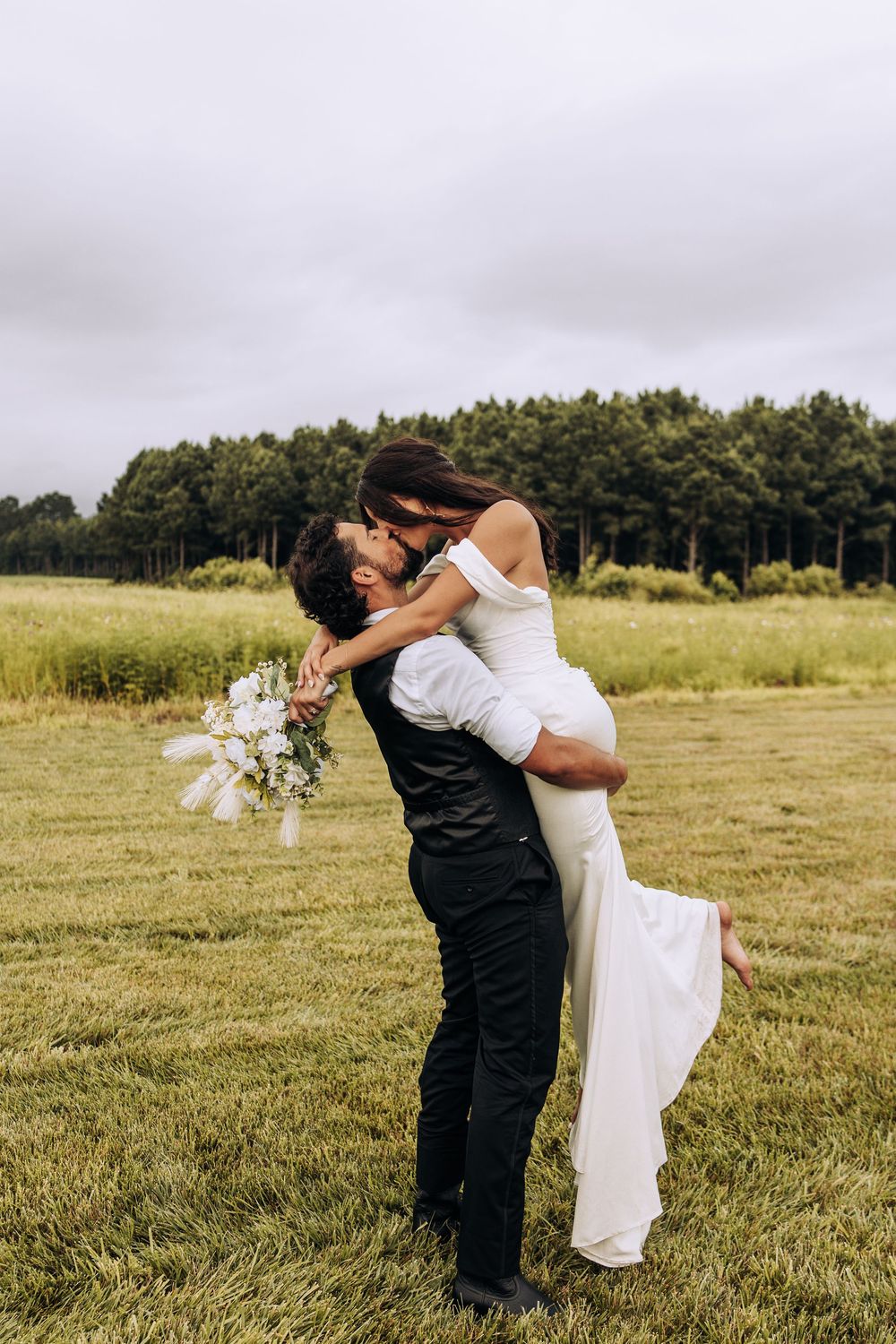 Groom lifts and kisses bride in white wedding dress outdoors in lush green field.
