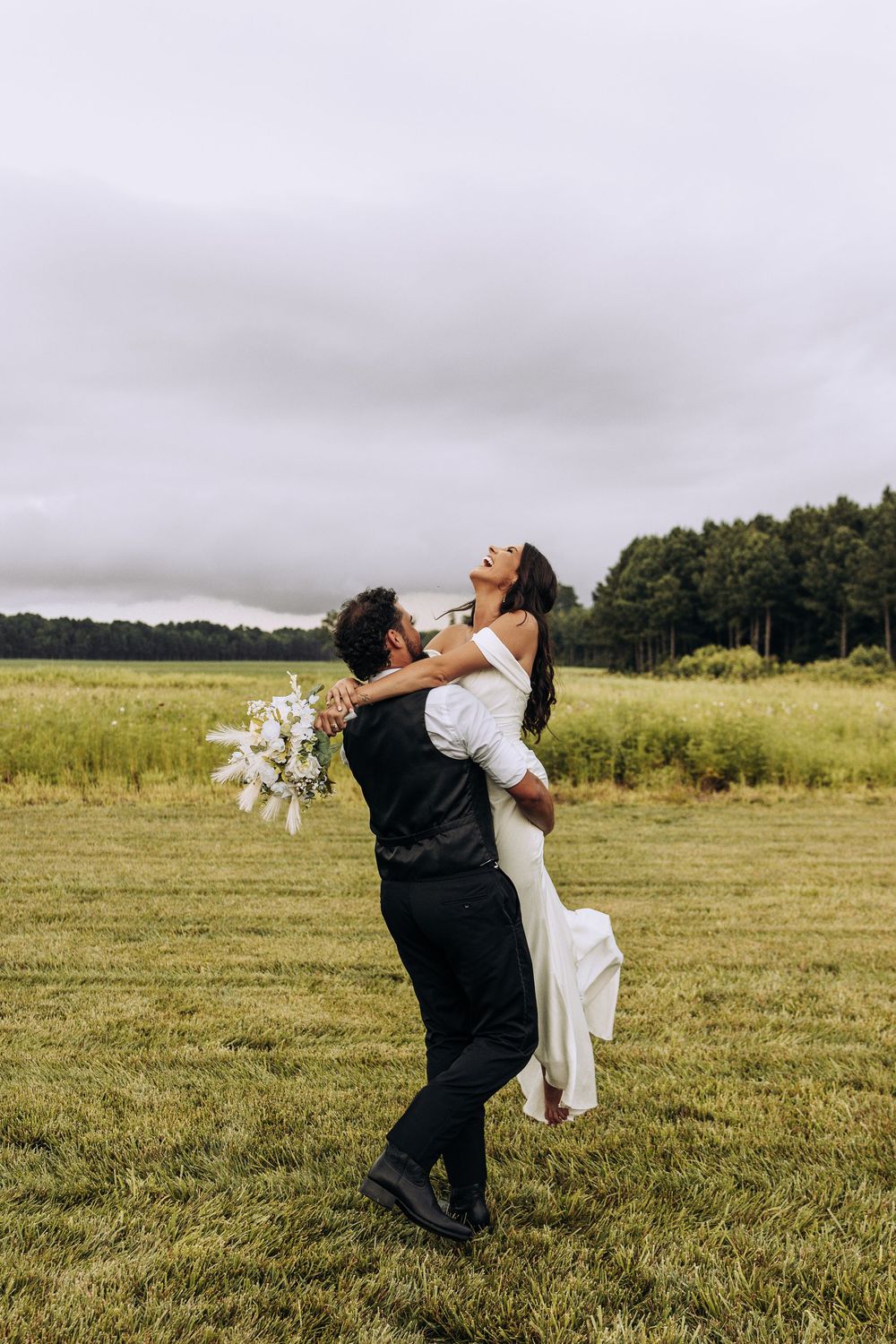 Joyful bride laughs as groom lifts her in white gown in scenic green meadow.