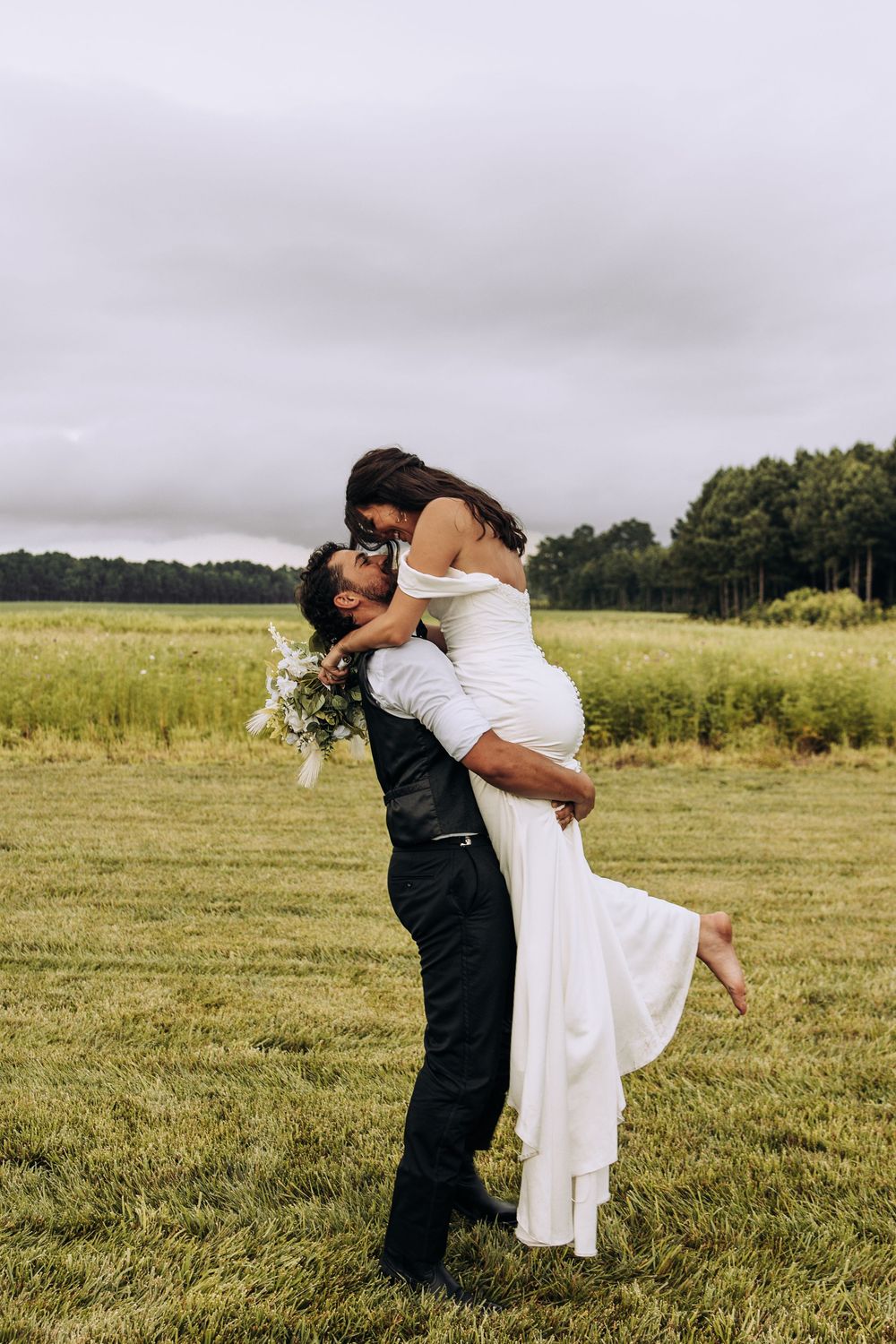 Groom lifts smiling bride in elegant white dress in a green outdoor field setting.