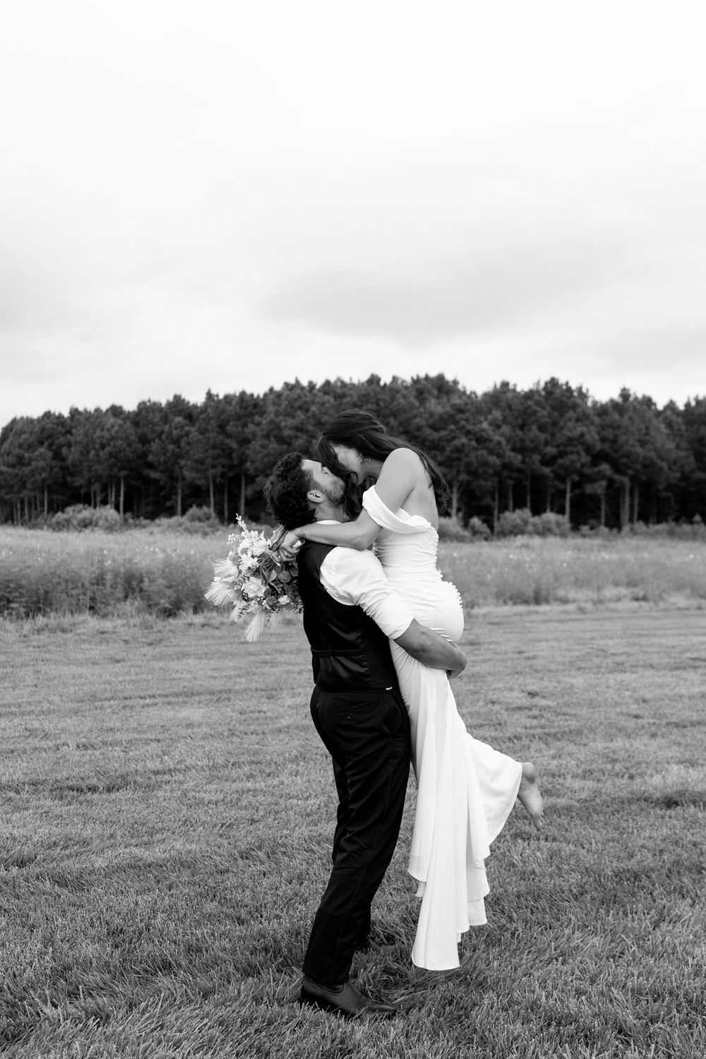 Black and white photo of groom lifting bride in white gown in open green field.