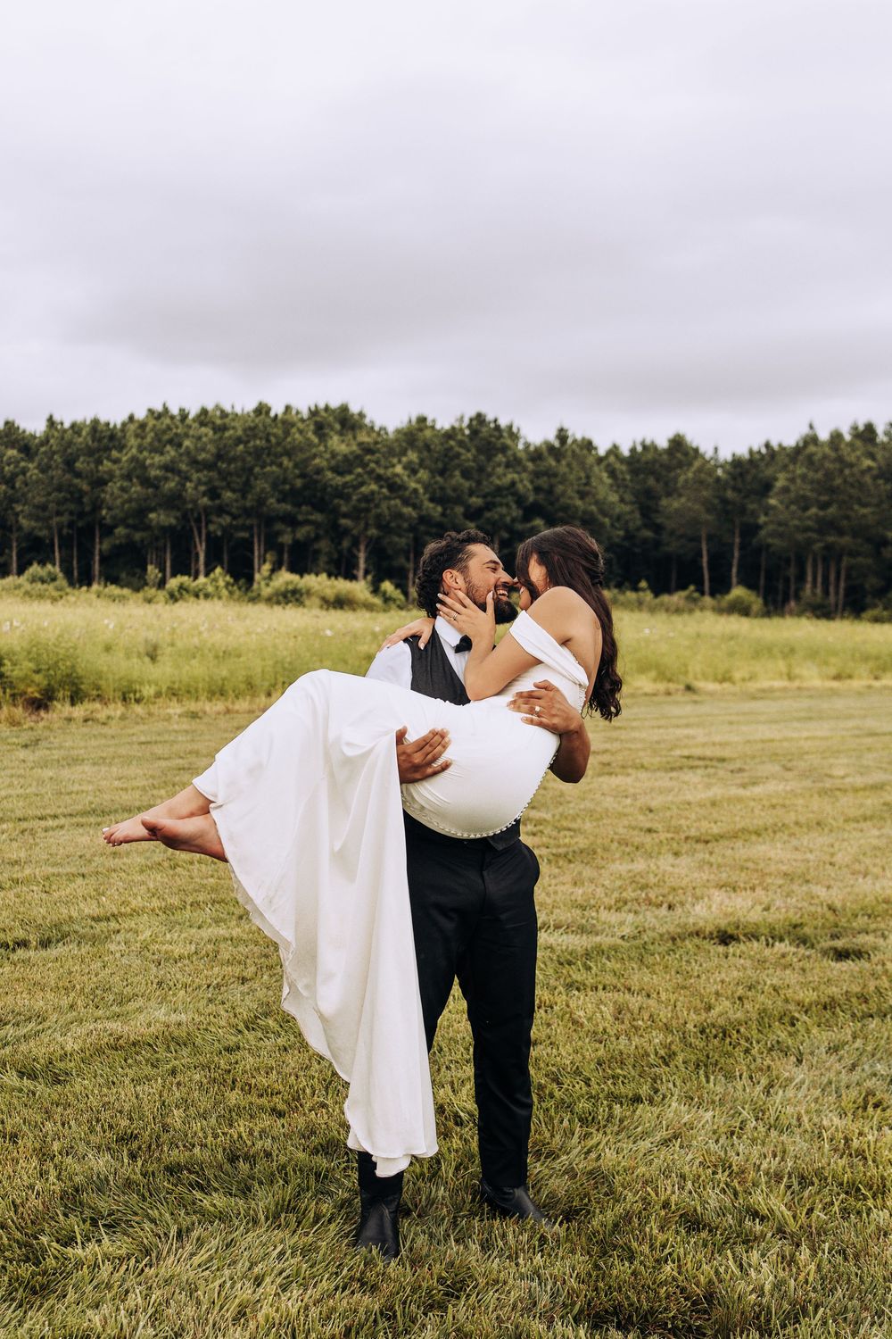 Groom carries barefoot bride in white dress in his arms across a green meadow.