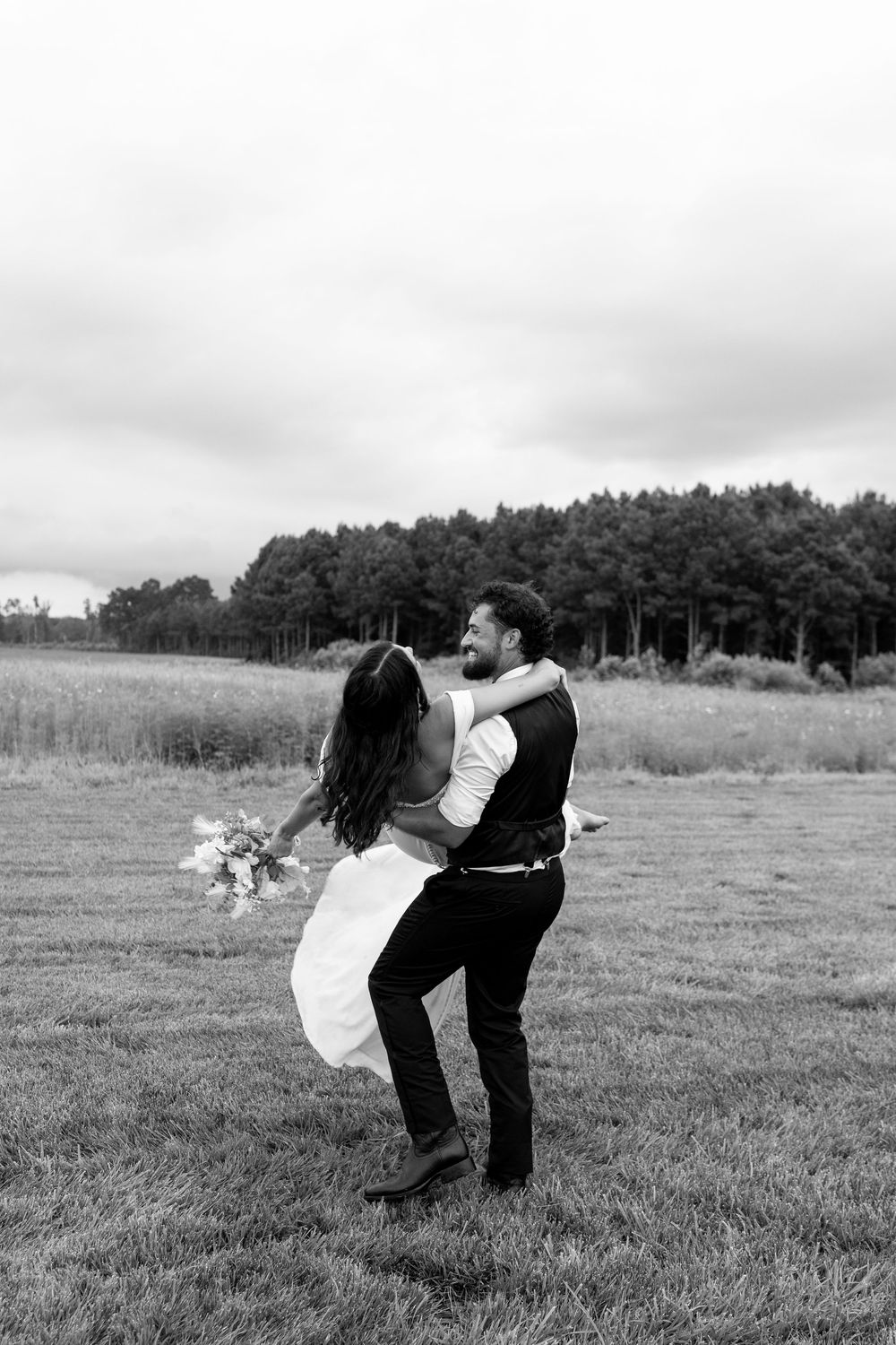 Black and white photo of groom spinning laughing bride in white dress in field.