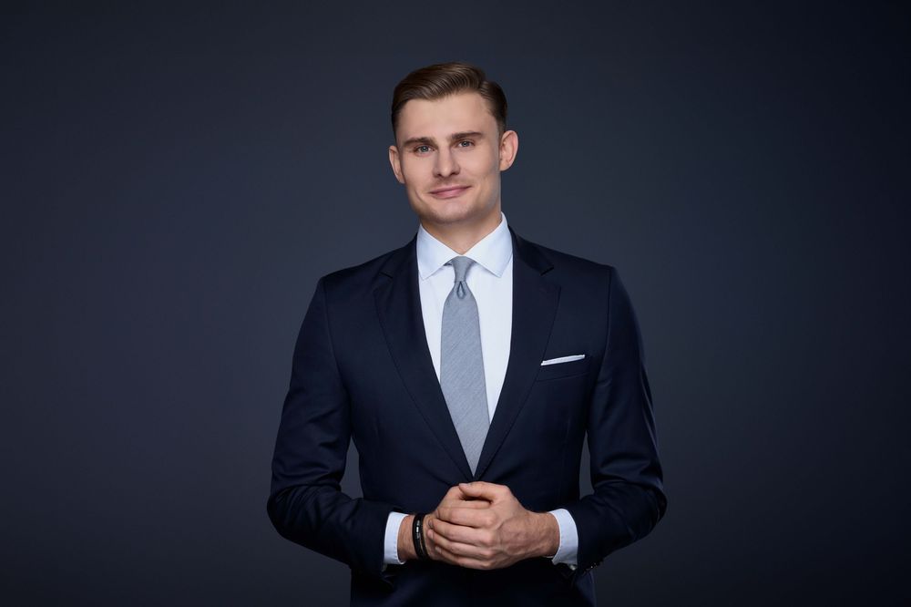 Professional headshot of a person in a dark suit and tie against a dramatic dark background.