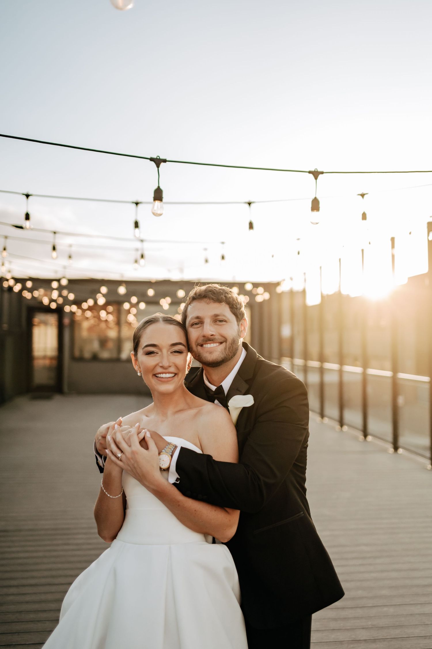 Bride and groom embrace on rooftop terrace at golden hour with string lights overhead.