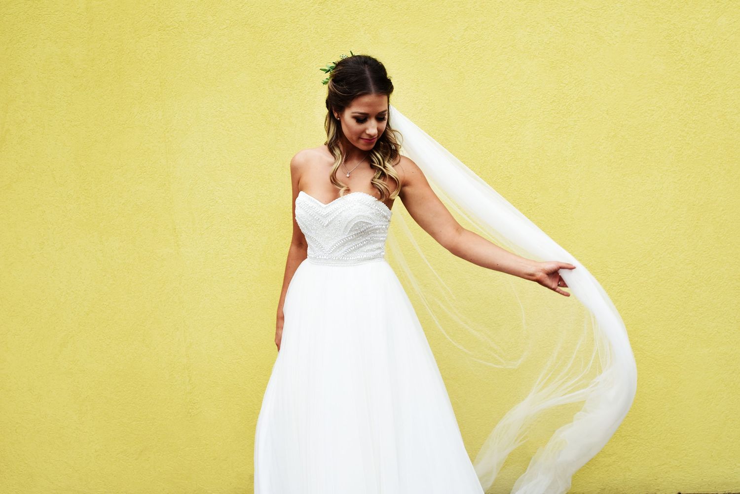 Mariée souriante tenant son voile sur mur jaune. Elle porte une robe bustier simple avec des perles brodées. Mariage intimiste à Montreal.