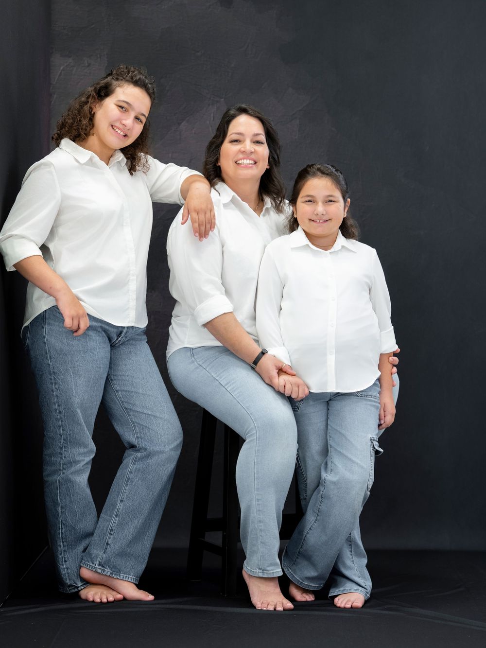Three women in matching white shirts and jeans pose together for a family portrait against a dark background.