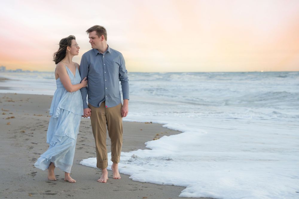 Couple holding hands walking barefoot on a sandy beach at sunset with ocean waves nearby.