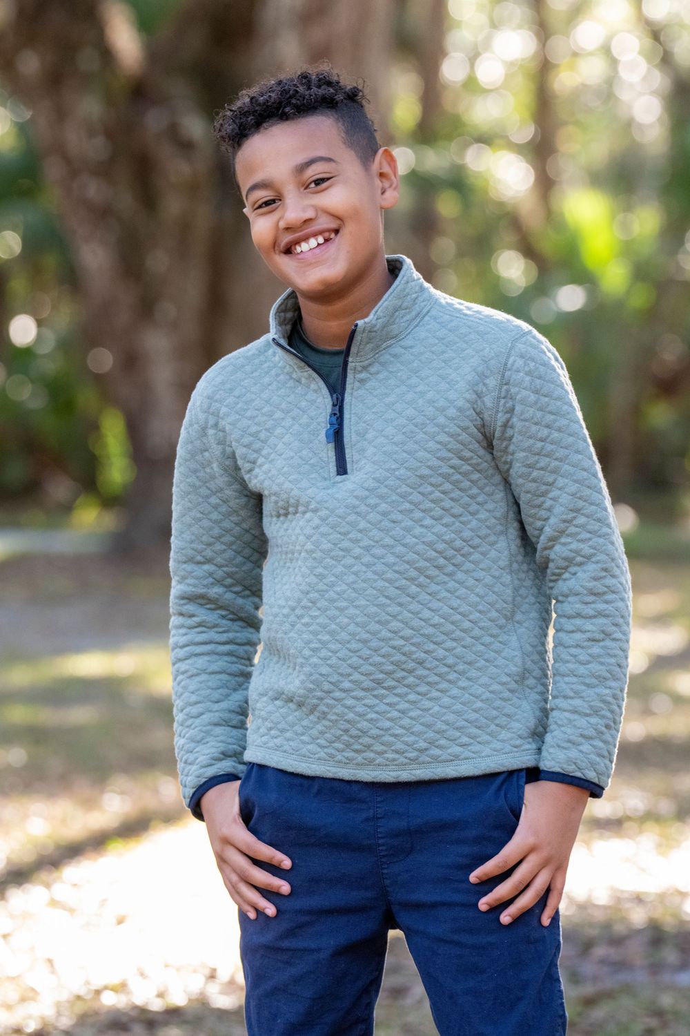 Young child wearing a gray quilted pullover and navy pants poses playfully in an outdoor setting.