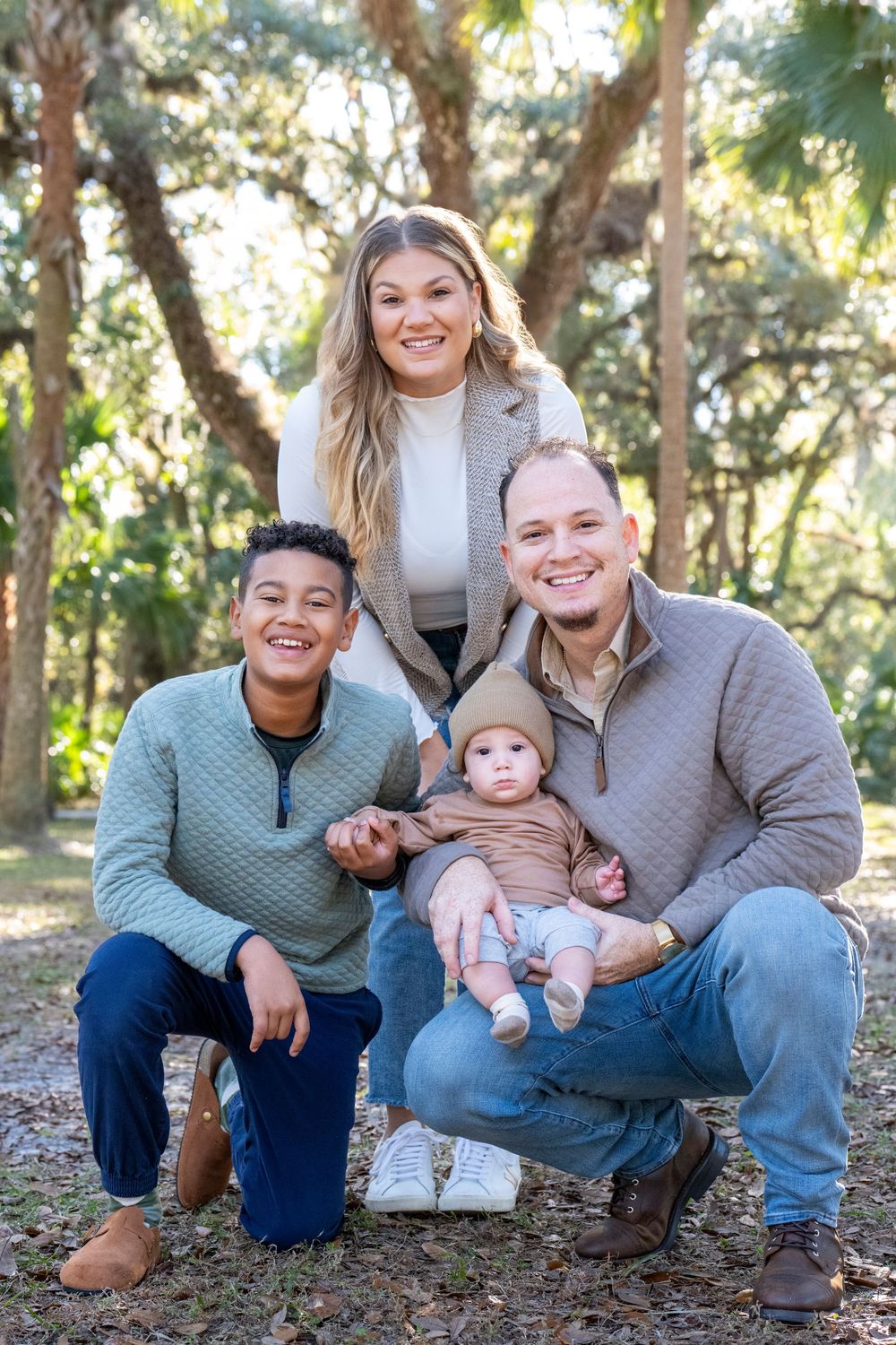 A family sitting together outdoors in a wooded area with fallen leaves on a sunny autumn day.