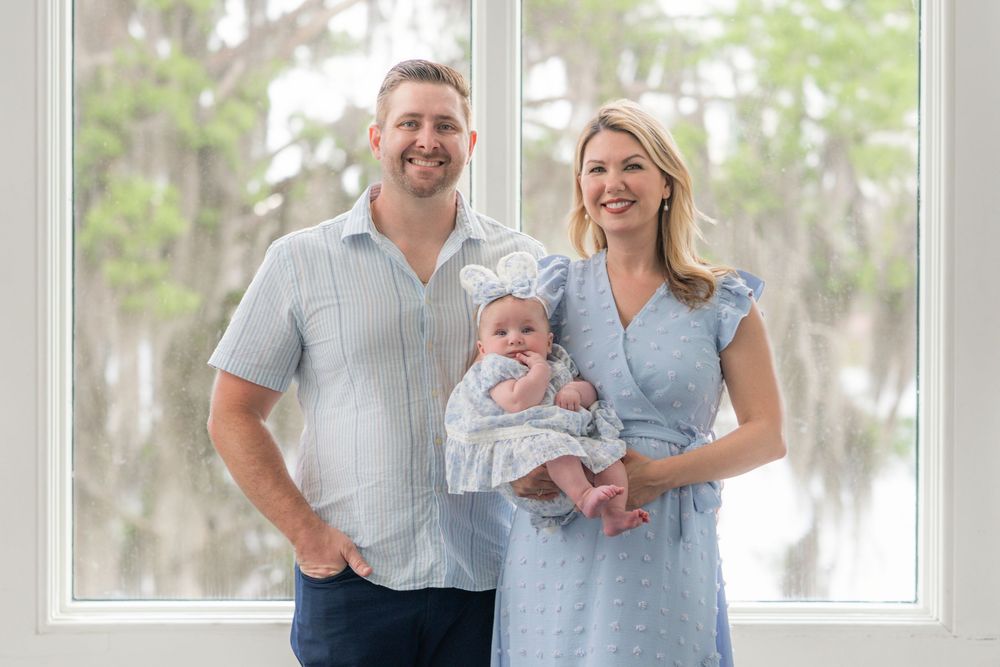 Family wearing coordinated blue and white outfits poses together for outdoor portrait session in front of mossy trees.
