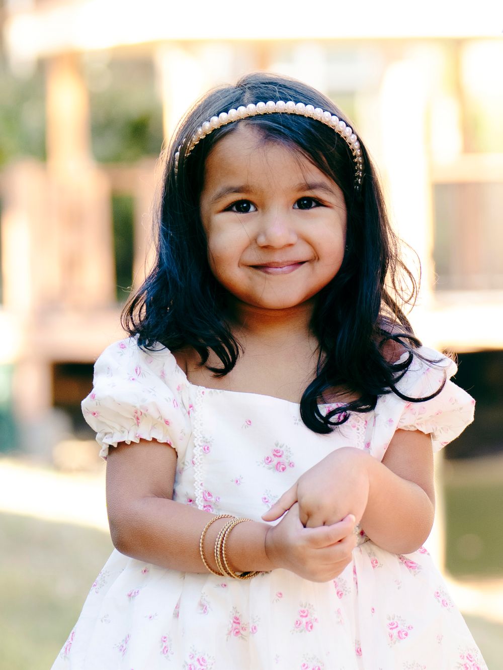A small child wearing a white floral dress and sparkly headband smiles warmly outdoors in natural lighting.