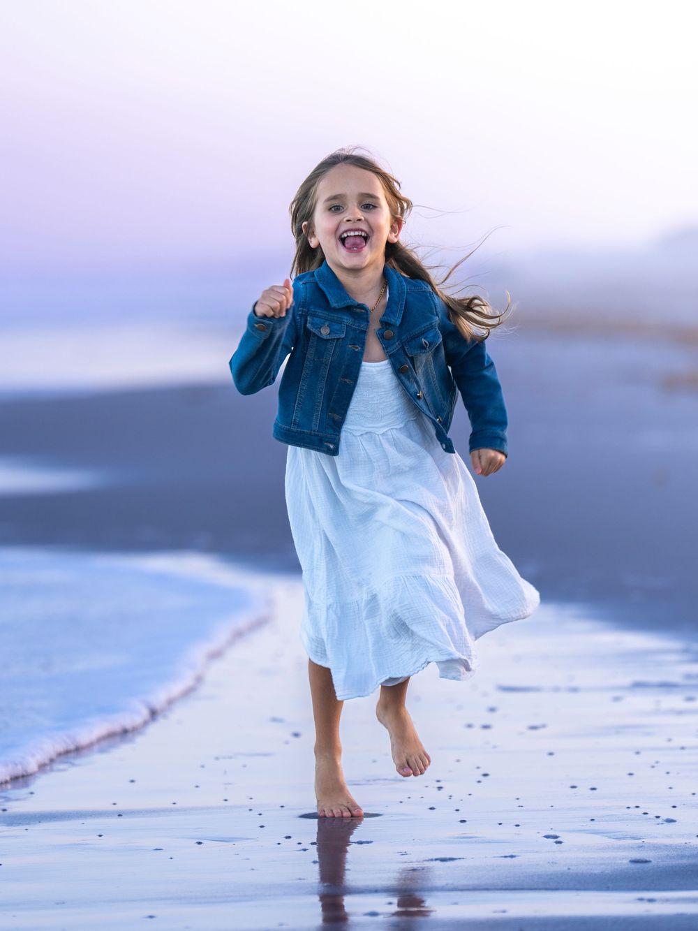 Young person in white dress and denim jacket joyfully running along the beach at sunset.