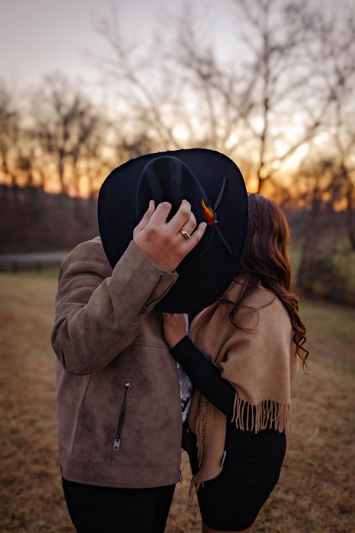 Couple sharing a kiss behind a cowboy hat in a golden field during golden hour Northern Virginia engagement photography.