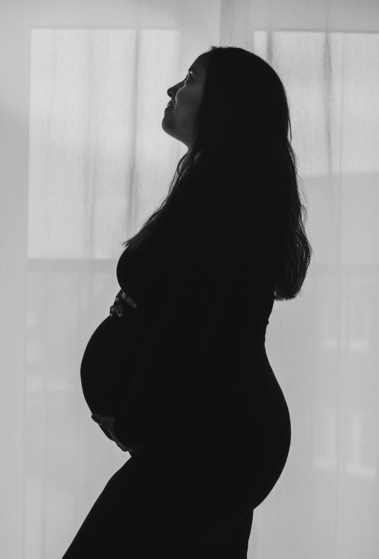A black and white silhouette of a pregnant woman standing in profile against a light window background.
