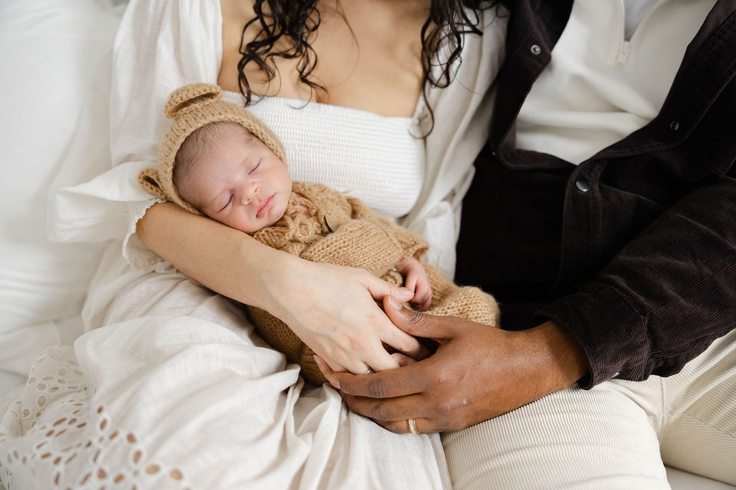 Parents sitting together on a bed holding their Newborn baby sleeping peacefully while bein baby during a relaxed in-home lifestyle session.