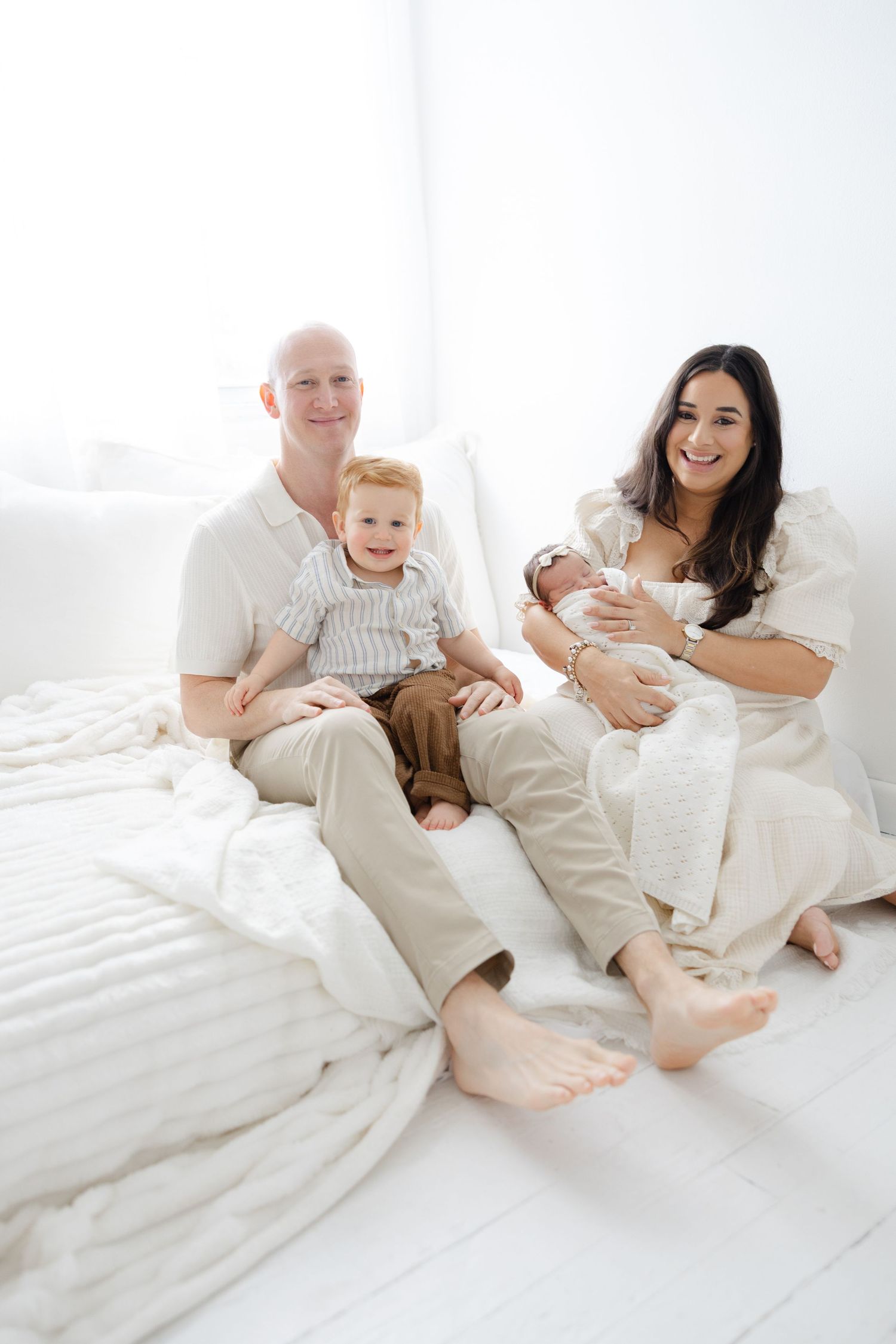 Parents sitting together on a bed holding their newborn baby during a relaxed in-home lifestyle session.