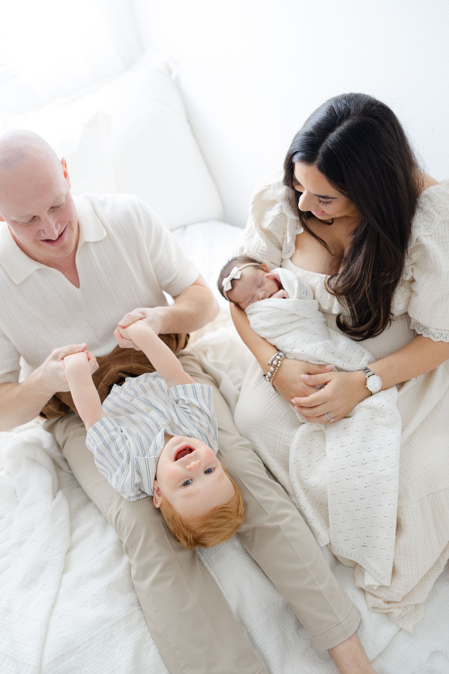 Lifestyle family photo with parents and toddler interacting with their newborn baby on a bed in natural light.