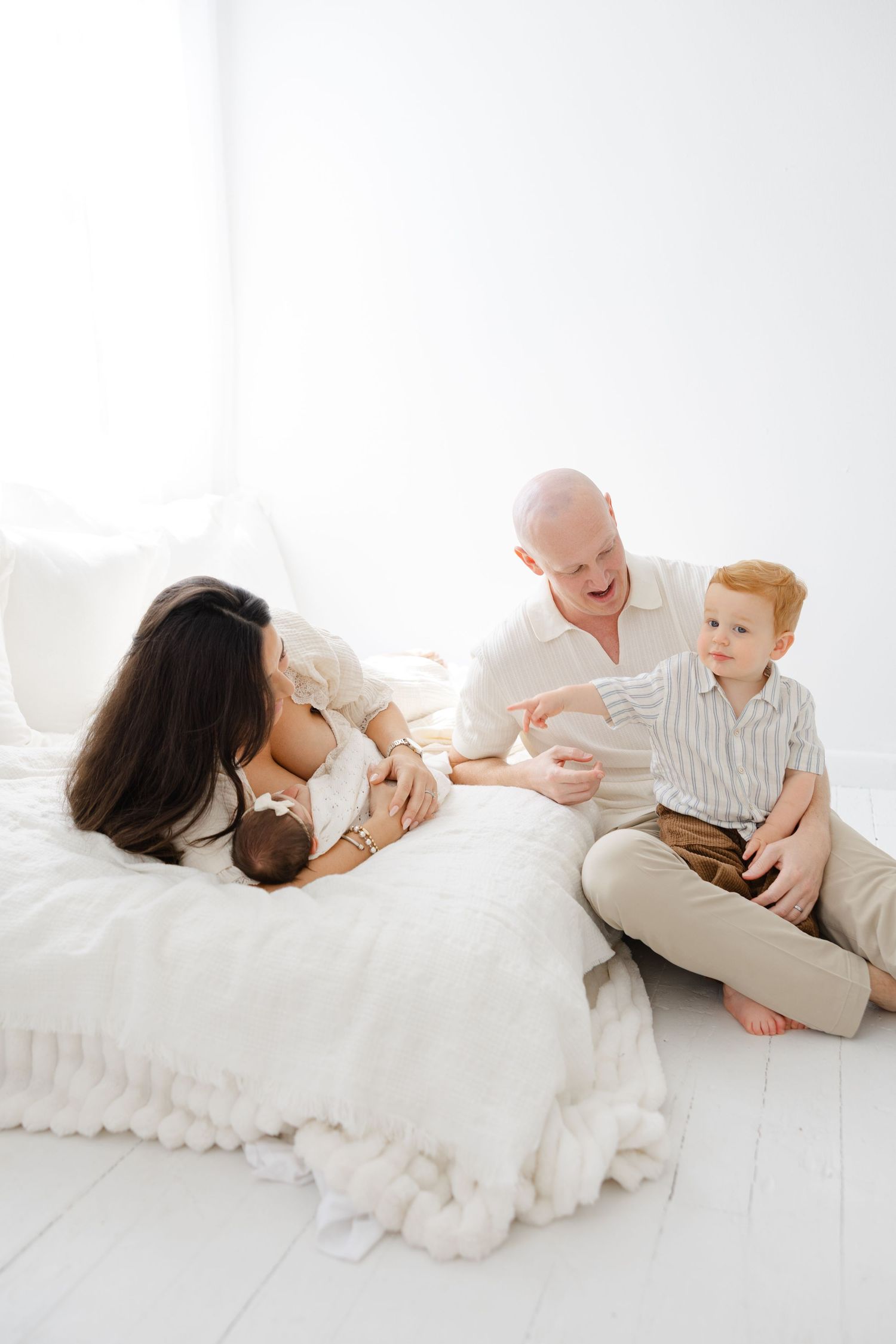 Parents sitting together on a bed holding their newborn baby in a bright, minimal lifestyle photography setting.