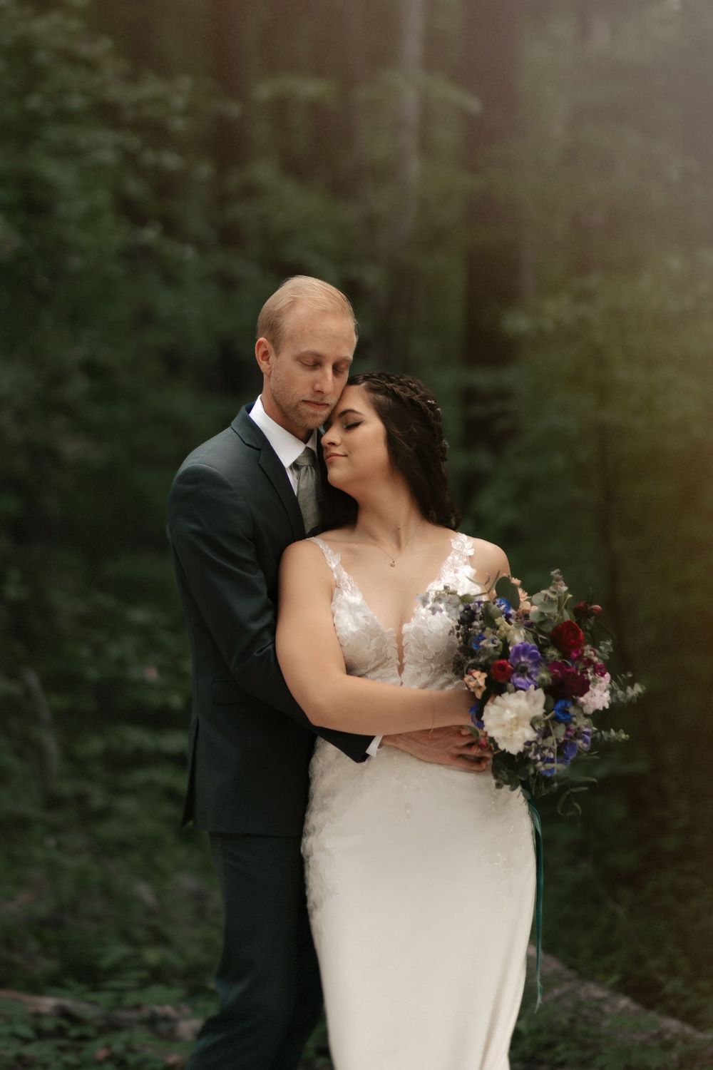 Romantic wedding couple embraces in a forest setting with a beautiful bouquet of purple and white flowers.