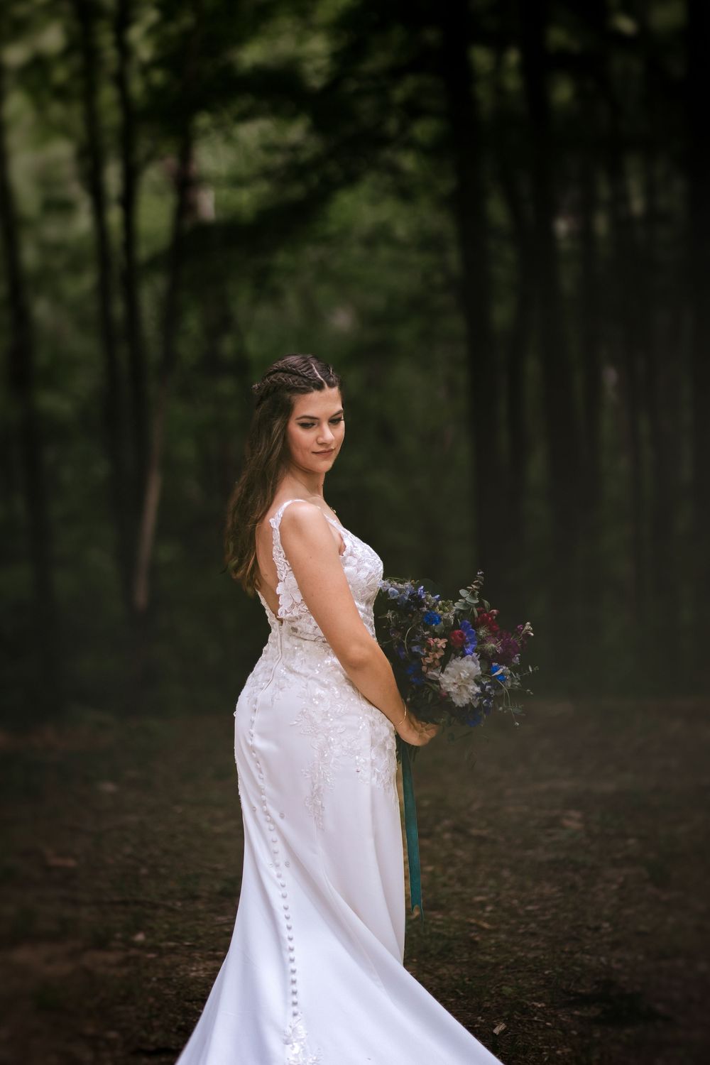 Bride in a flowing white lace gown poses elegantly in a wooded setting with a colorful bouquet.