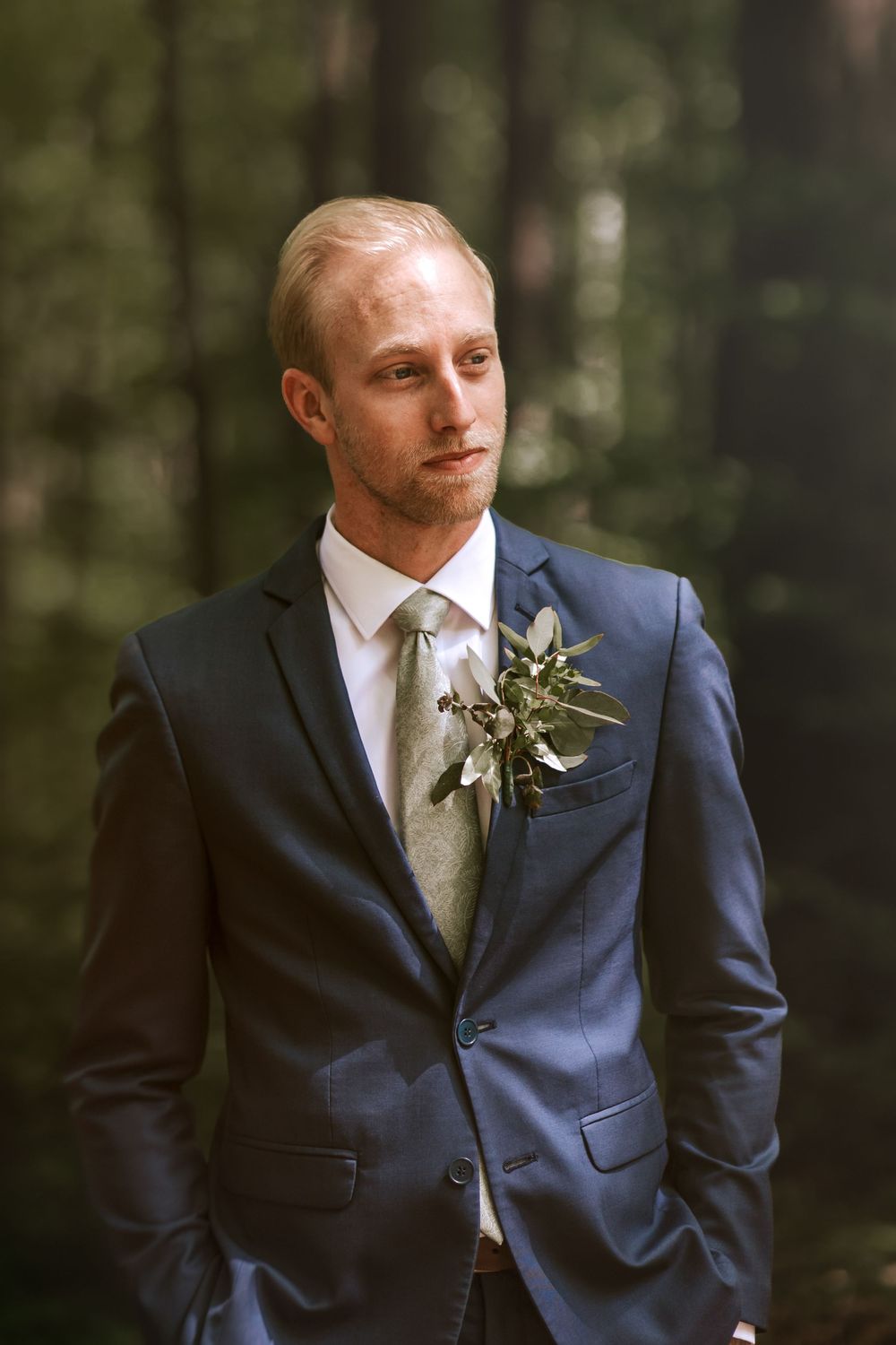 Groom in navy suit with sage tie poses thoughtfully in a forest setting.