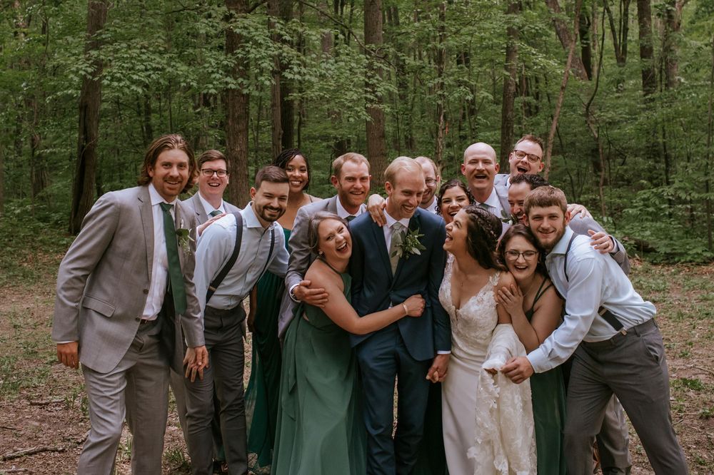Wedding party poses together in coordinated navy and grey attire against forest backdrop.